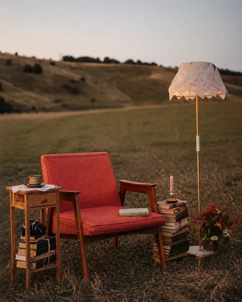 Червоне крісло, столик із чашкою, стоси книжок і торшер посеред поля на заході сонця — фото власної локації